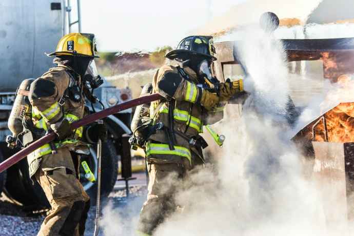 Two firefighters in protective gear spraying water from a hose onto a fire